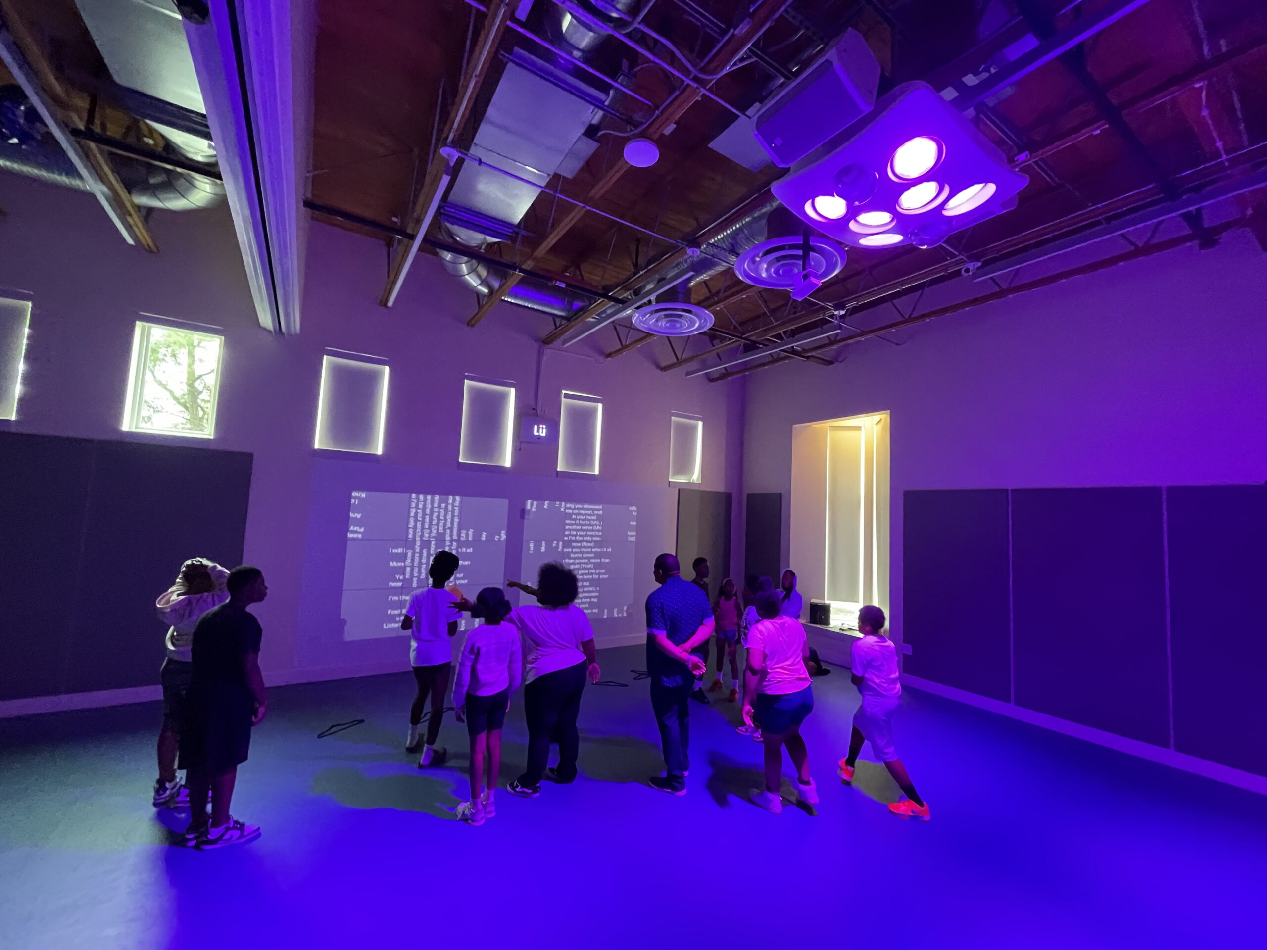 Students and a staff member gathered in the multipurpose room, lit purple by the Lü light rig, facing a word-based Lü app projected on the wall.