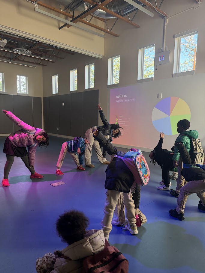 Students stretching during a Lü movement activity as a results wheel is projected on the wall of the daylit multipurpose room.