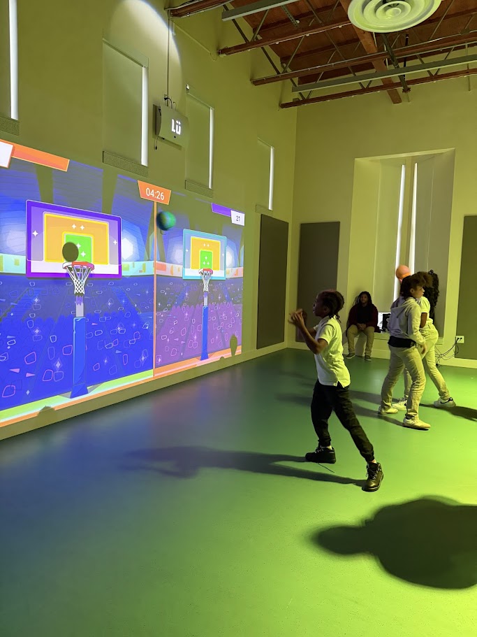 A student throwing a ball toward a Lü basketball game projected on the wall of the multipurpose room.