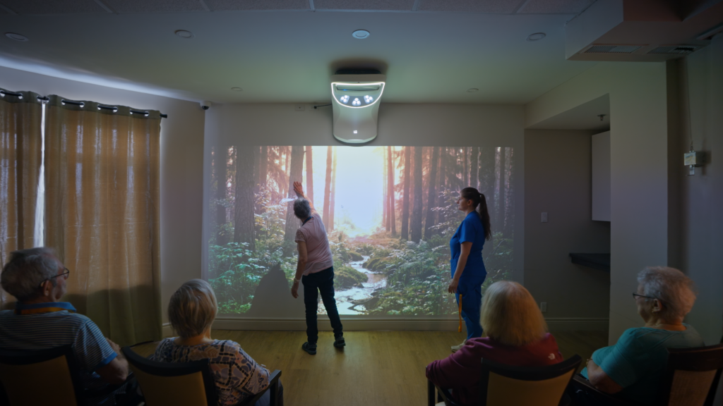 Une résidente tend la main vers une projection de forêt pendant qu'une préposée l'accompagne et que d'autres résidents observent depuis leur fauteuil