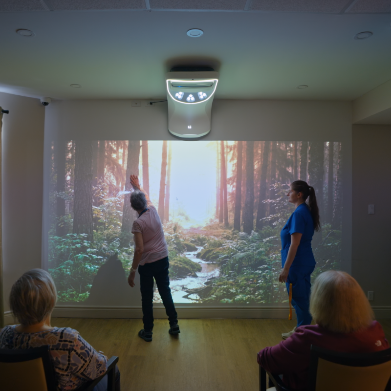 Une résidente tend la main vers une projection de forêt pendant qu'une préposée l'accompagne et que d'autres résidents observent depuis leur fauteuil