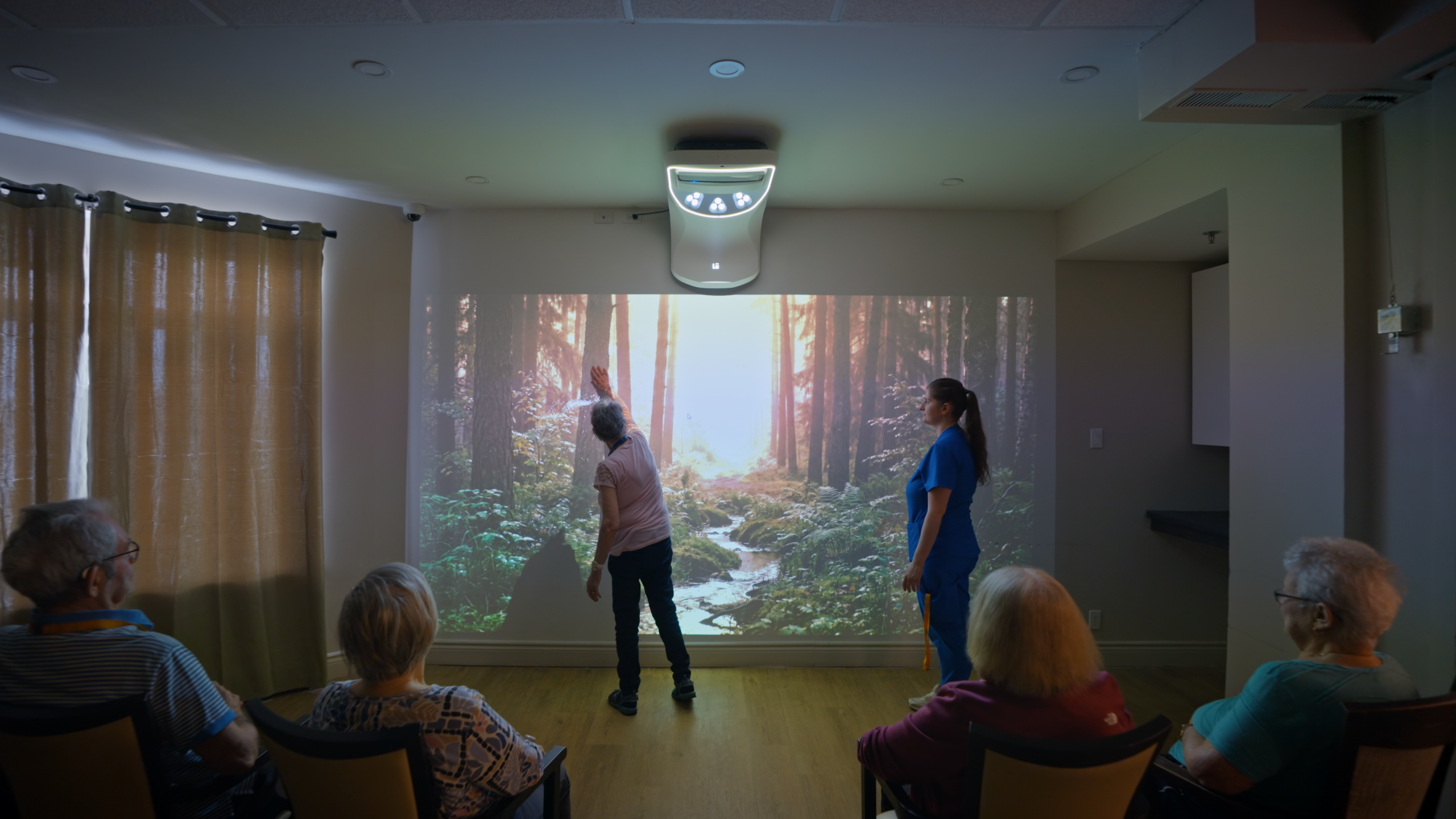Une résidente tend la main vers une projection de forêt pendant qu'une préposée l'accompagne et que d'autres résidents observent depuis leur fauteuil