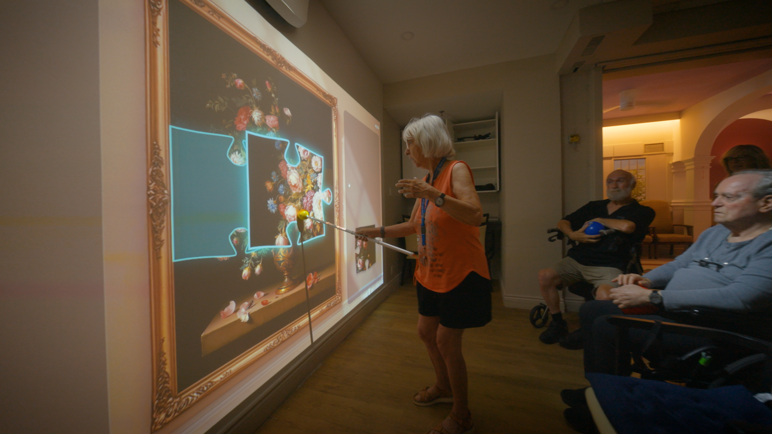 A resident places a piece of a projected floral painting puzzle while other residents watch from the side of the room