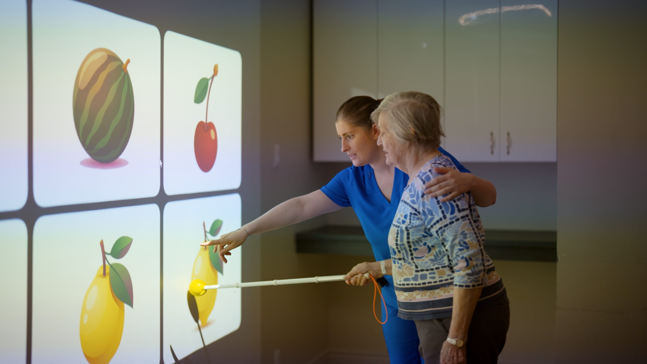 A caregiver guides a resident as she points to projected images of fruit with an extended pointer
