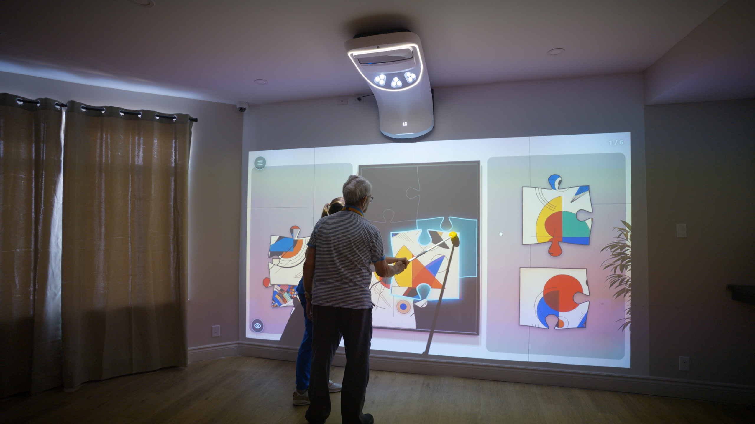 A resident uses an extended pointer to move pieces of a projected art puzzle, with a caregiver standing beside him